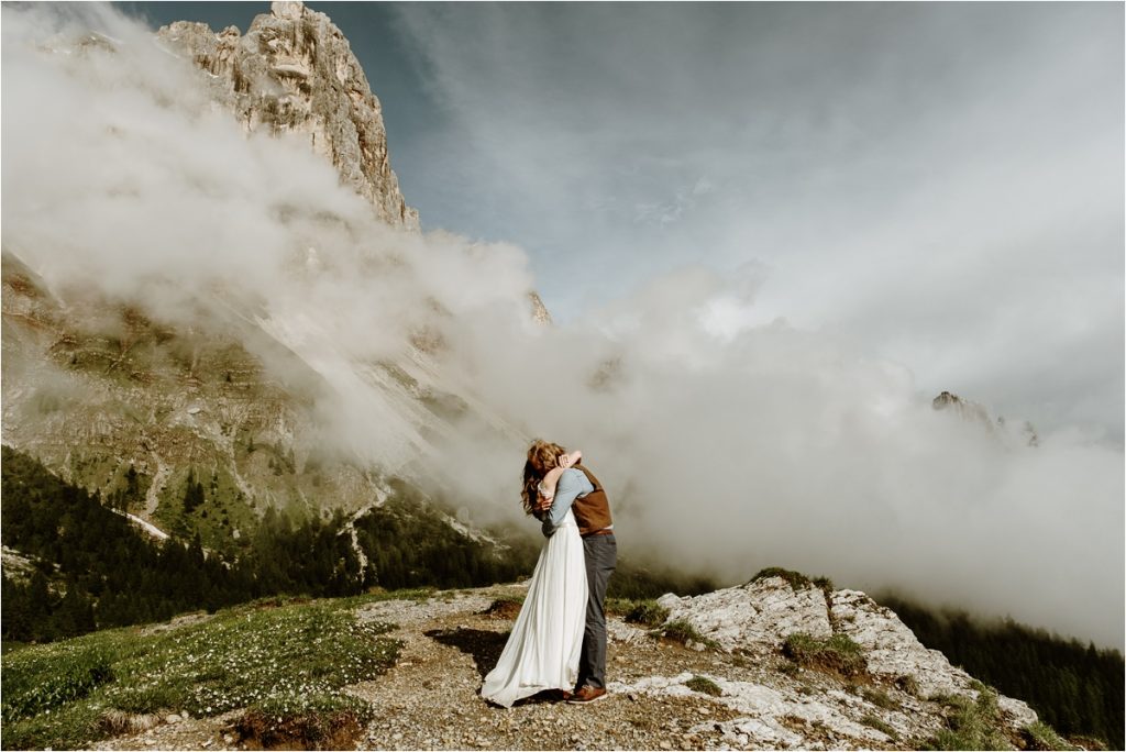 Elopement in the Italian Alps couple embraces on the top of a mountain with clouds all around them. Photo by Wild Connections Photography