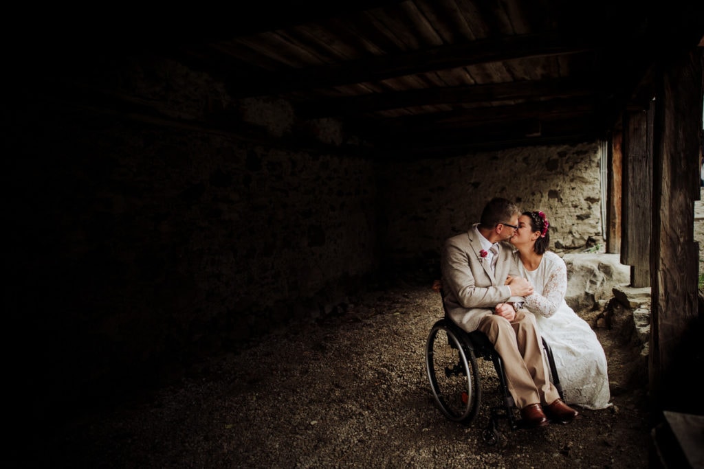 Bride and groom in a barn, groom is in a wheelchair and bride is crouching down to kiss him